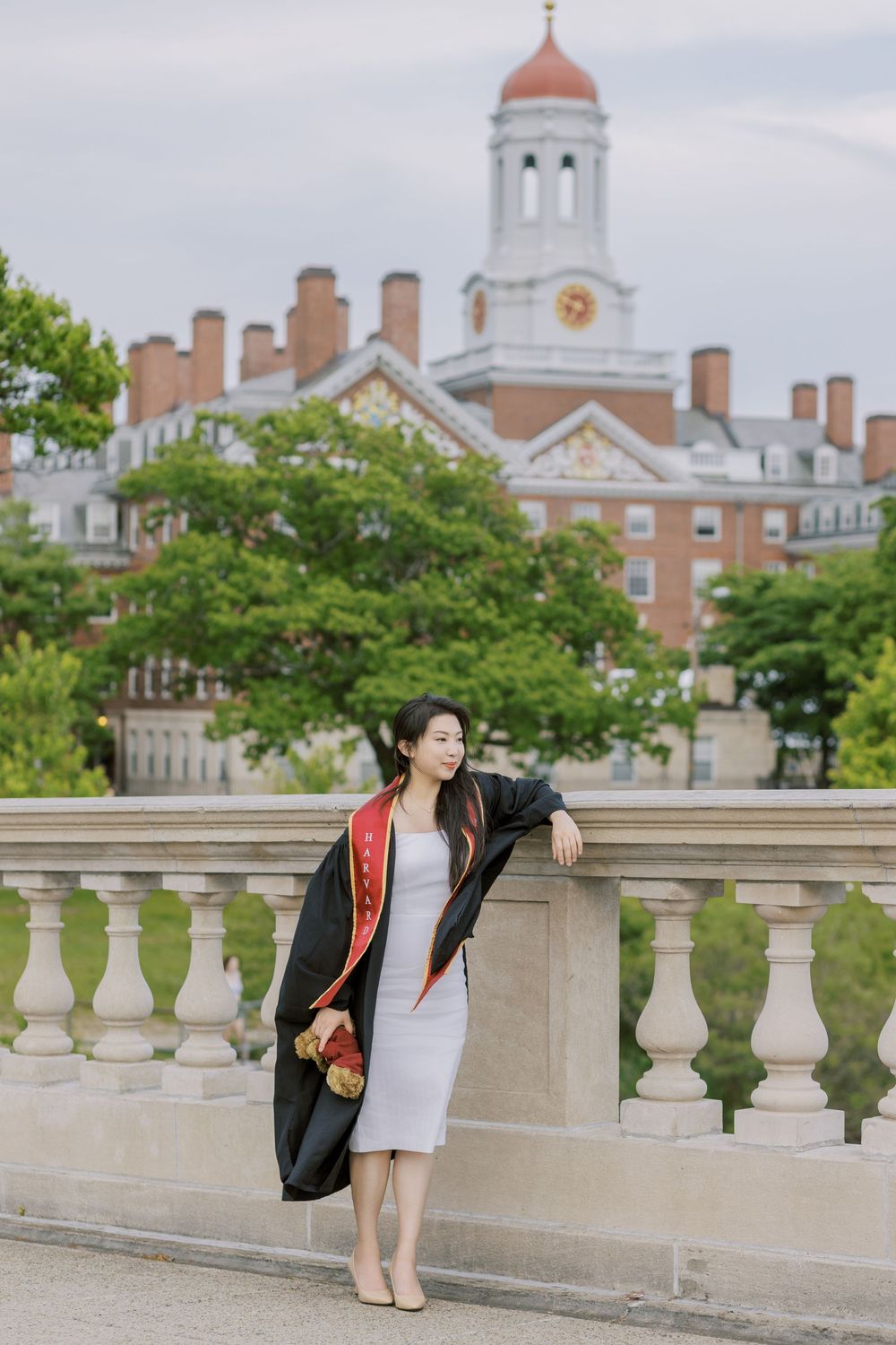 A young woman in Harvard graduation regalia poses on John W. Weeks Bridge with a historic brick university building in the background.