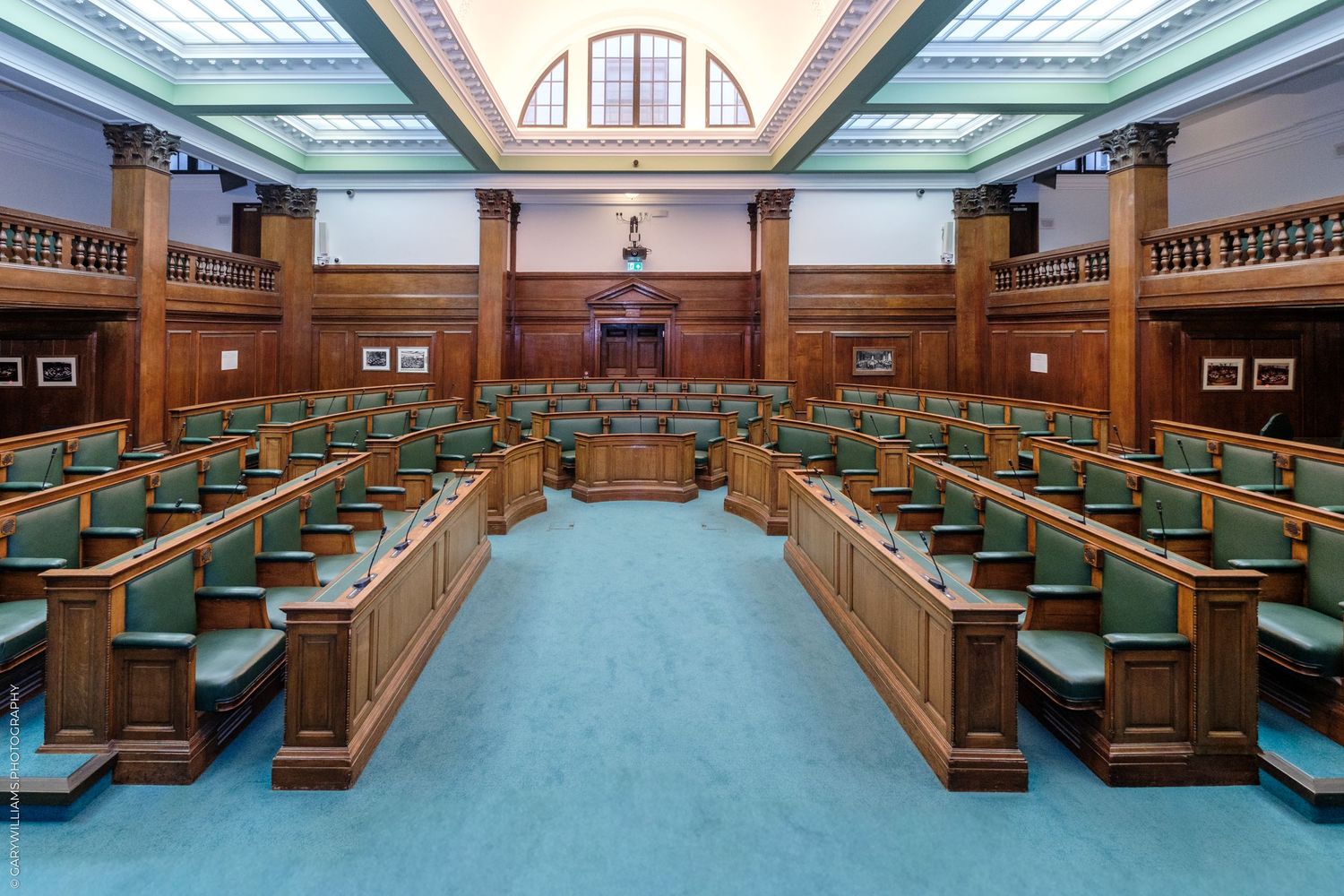Historic legislative chamber with green upholstered wooden benches, teal carpet, and ornate wooden paneling under arched windows.
