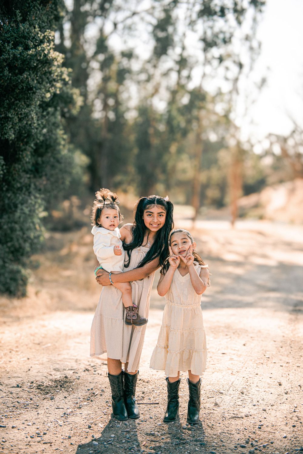 A woman in a cream dress and boots holds a baby while a young girl poses beside her on a sunny dirt path.