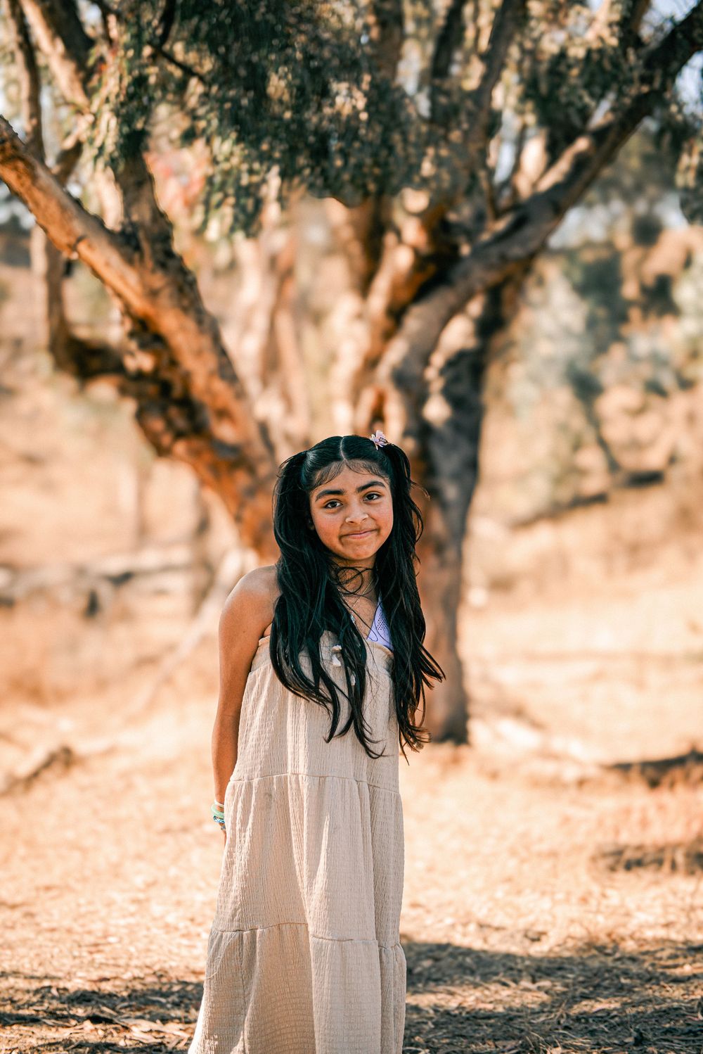 A young girl with long black hair in a white dress stands outdoors near a large tree in a sun-drenched natural setting.