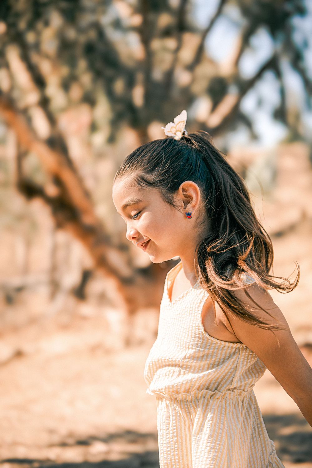 Young girl with flower hair clip in white dress smiling outdoors in a sun-drenched desert landscape.