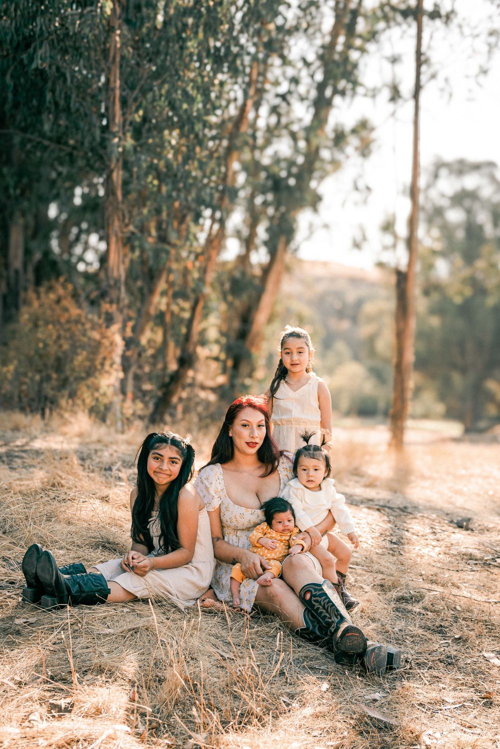 A mother sits outdoors with three young daughters on dry grass surrounded by tall trees in warm sunlight.