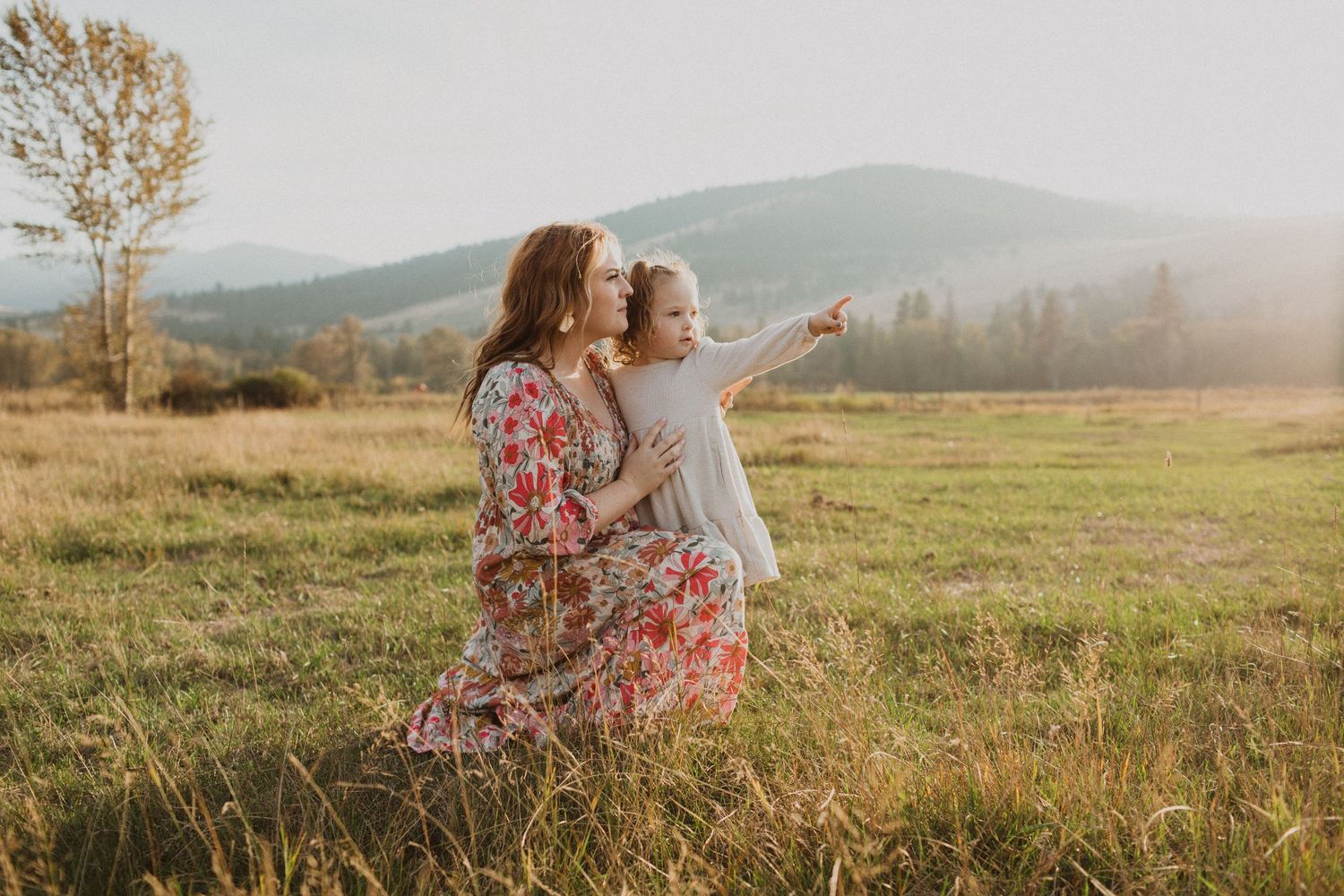 A mother and young daughter sit together in a sunlit meadow with mountain views, as the child points into the distance.