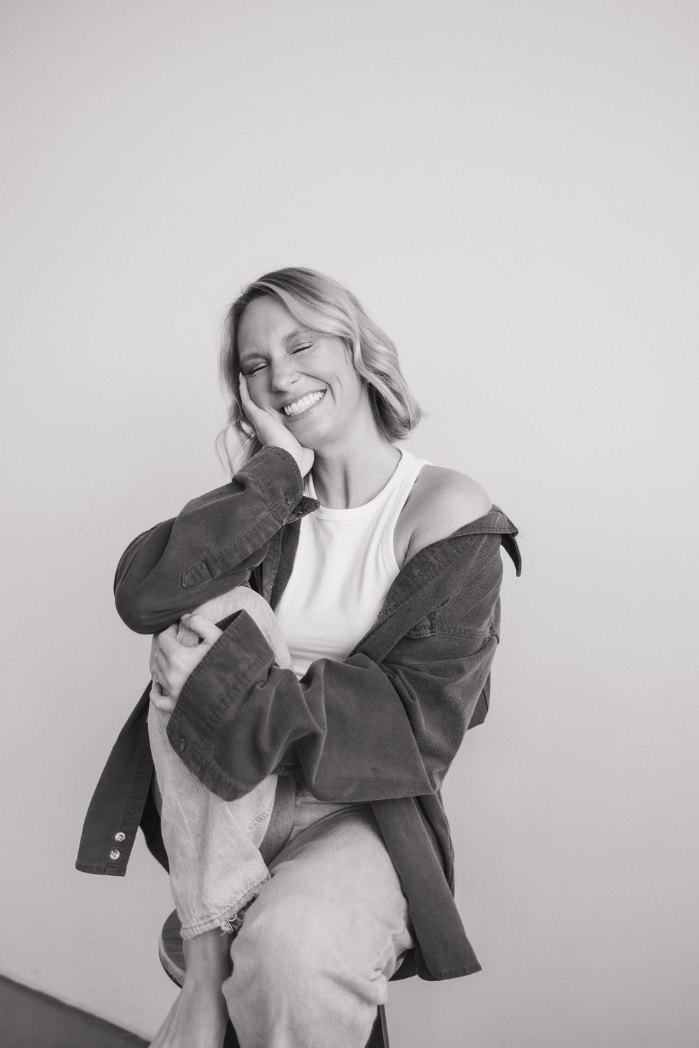 Smiling blonde woman in black and white photo wearing oversized jacket and white top, hand on cheek.