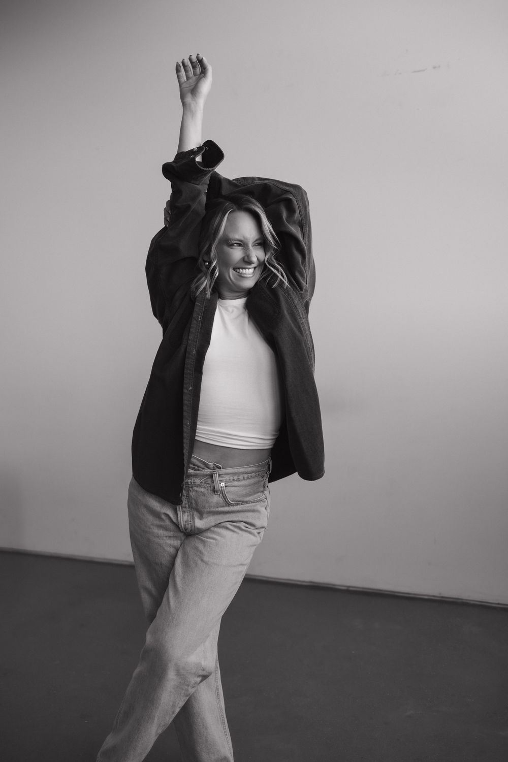 Black and white photo of a laughing blonde woman in jeans raising her arm while wearing an oversized jacket.