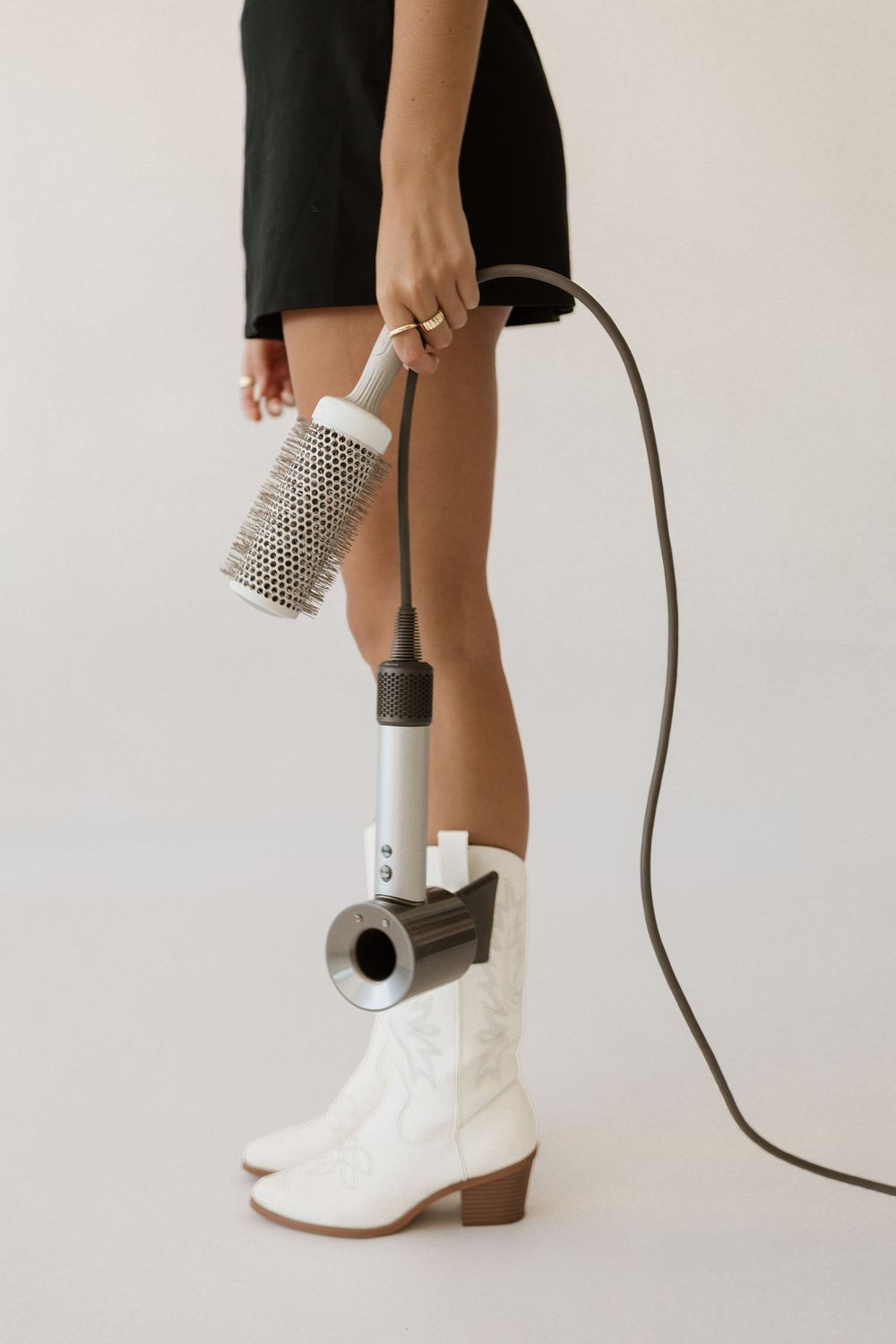 Woman in black dress and white cowboy boots holding a hair dryer with a round brush attachment.