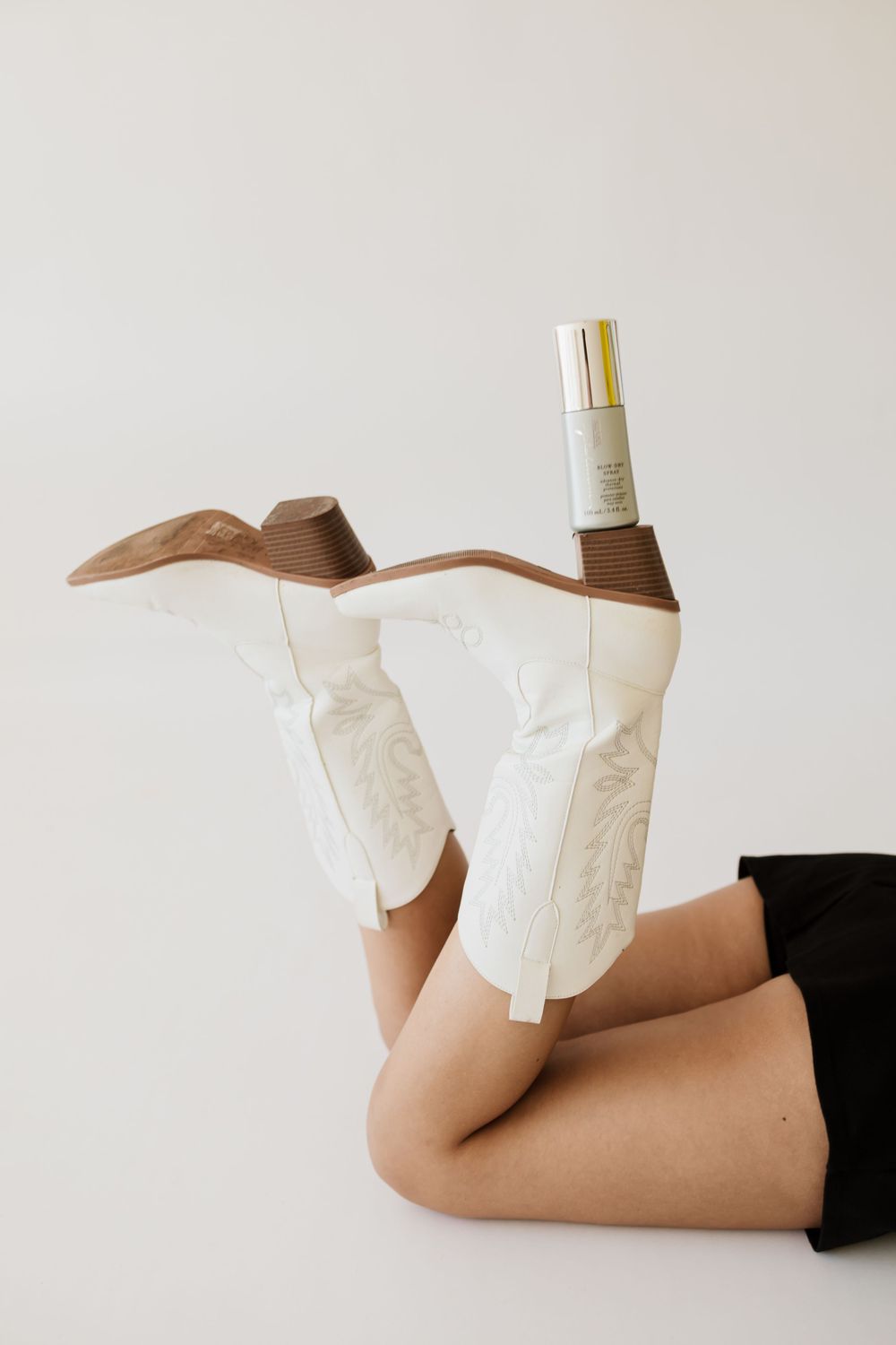 Woman lying down with legs raised wearing white cowboy boots, balancing a hair product on her foot.