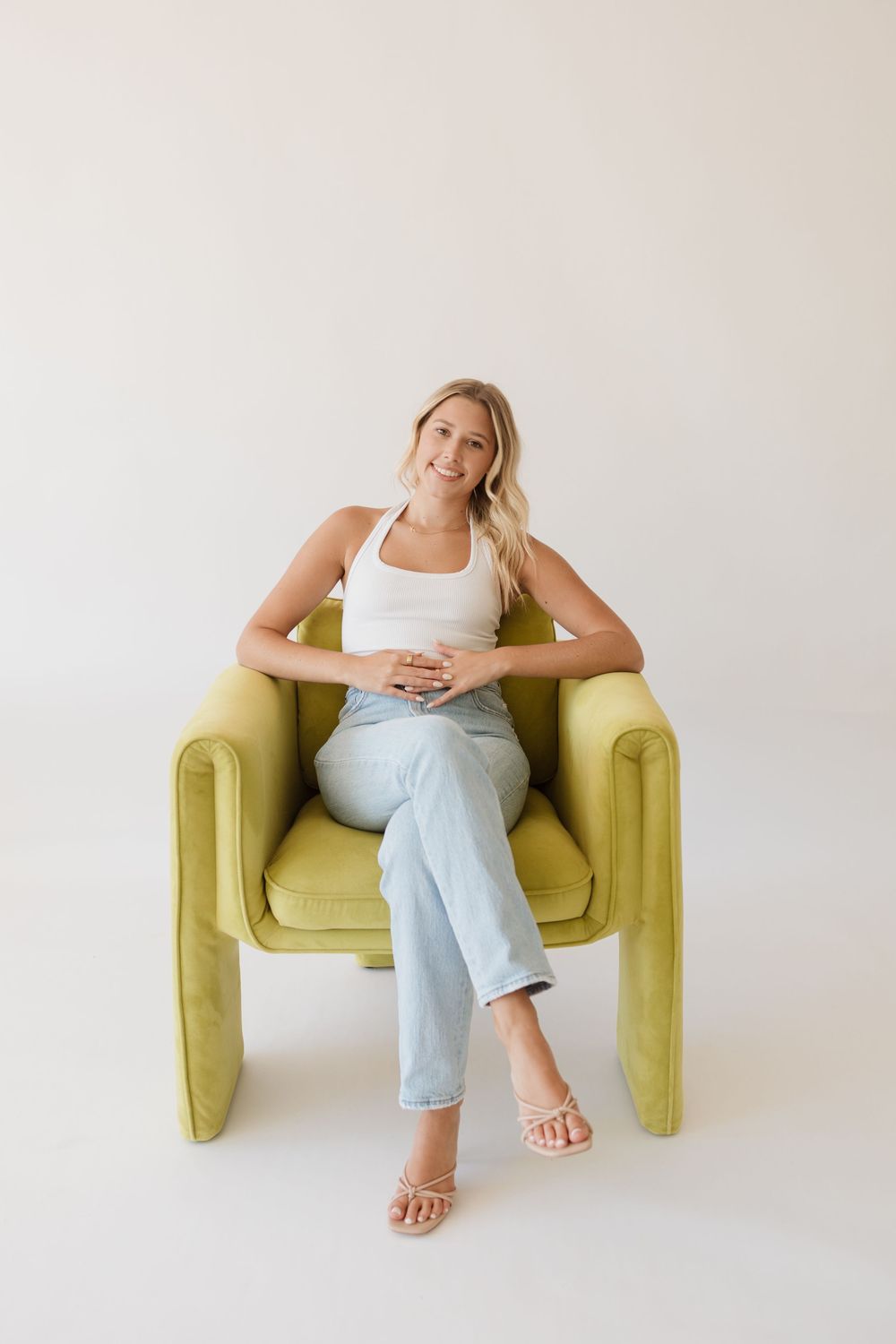 Smiling blonde woman in white tank top and light jeans sitting in a chartreuse green armchair.