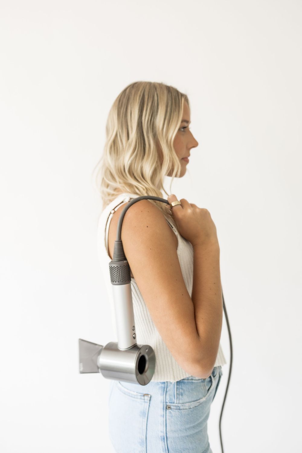 Blonde woman in white tank top and jeans holding a Dyson hair dryer against a white background.