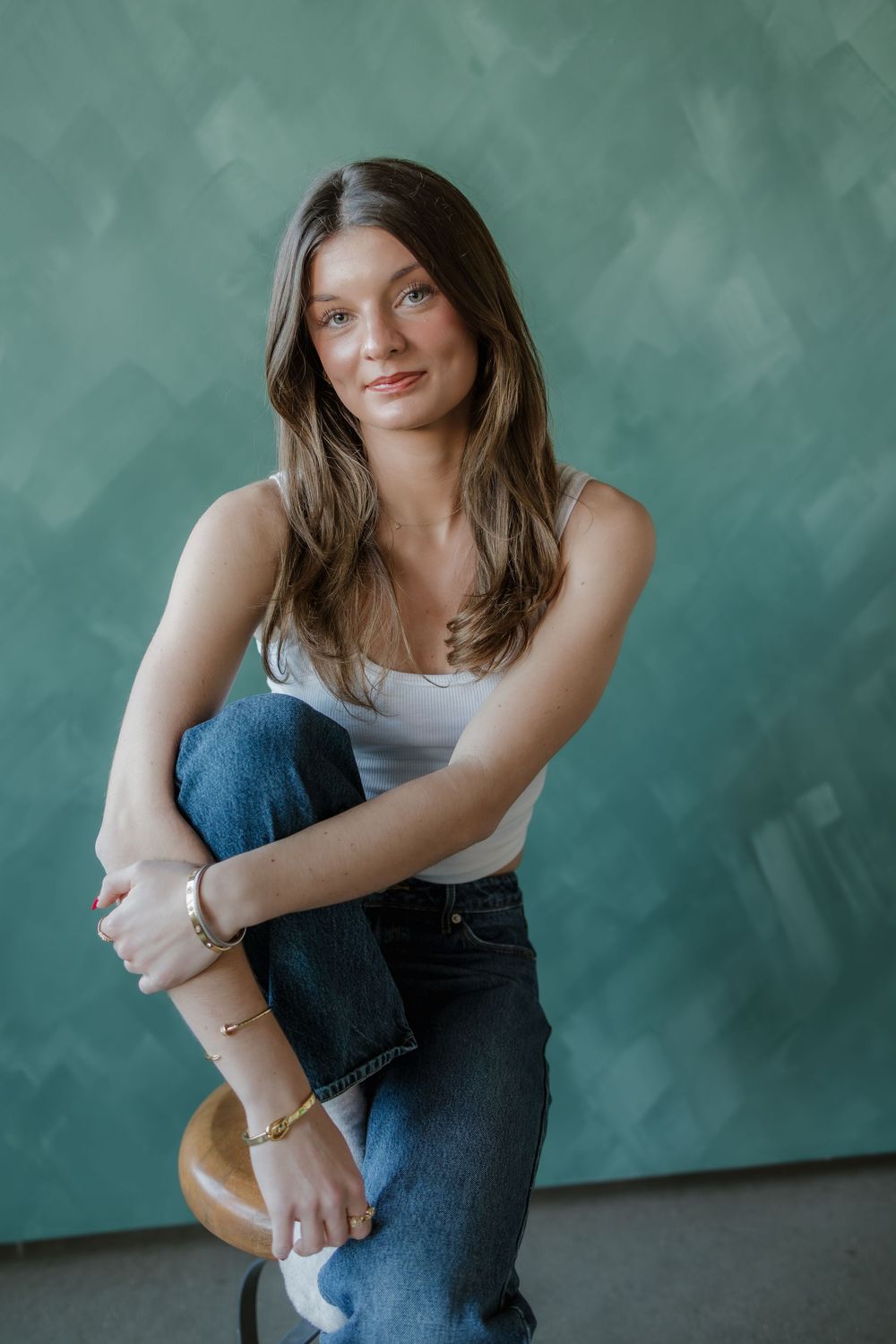 Young brunette woman sitting on stool in white tank top and jeans.
