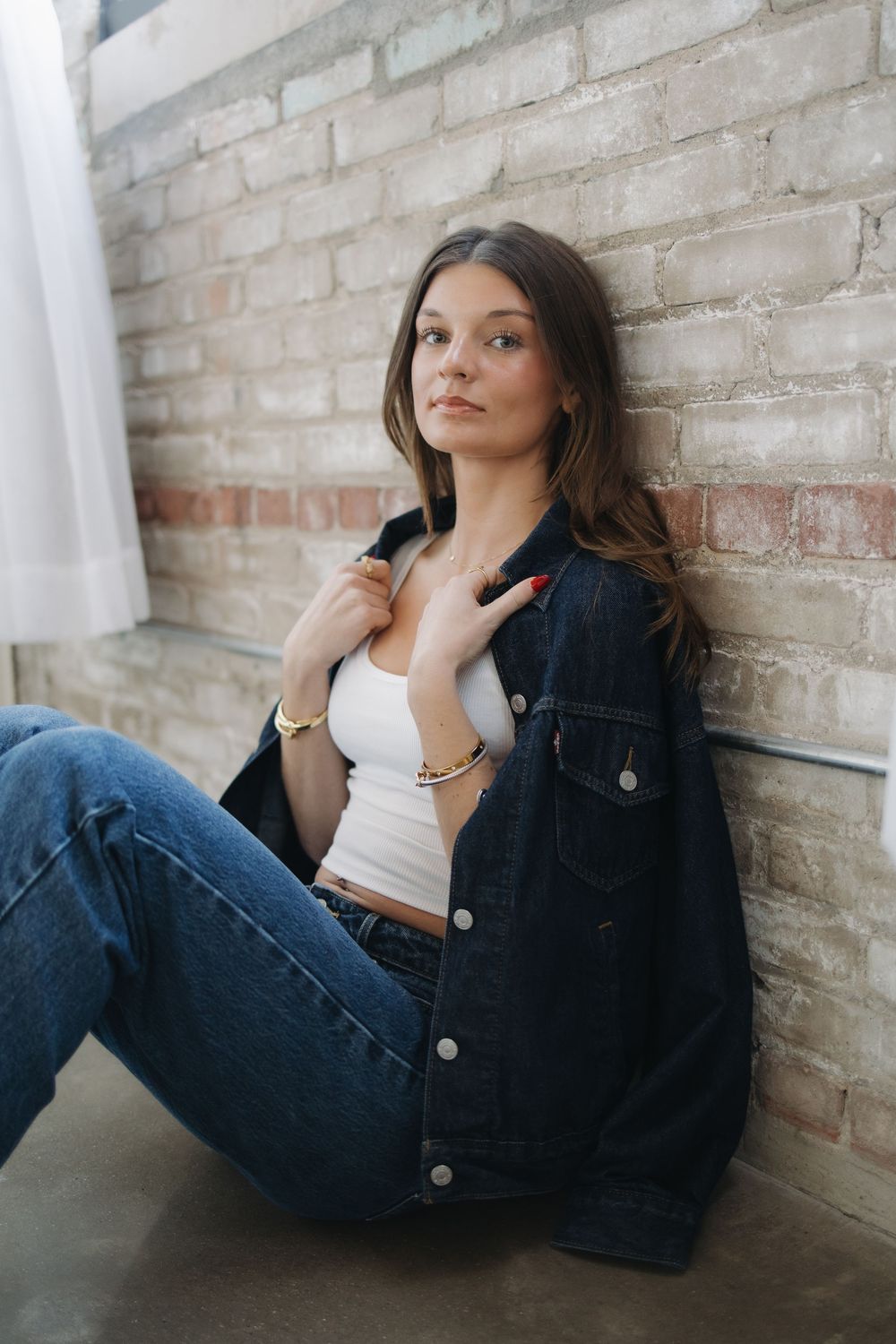 Young woman in denim jacket and jeans leaning against brick wall indoors.