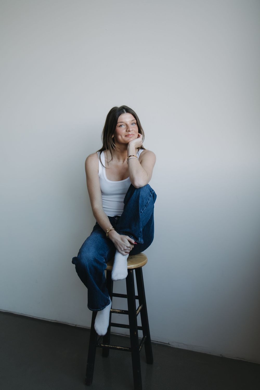 Young woman in white tank top and blue jeans sitting relaxed on a wooden stool against a light gray wall.