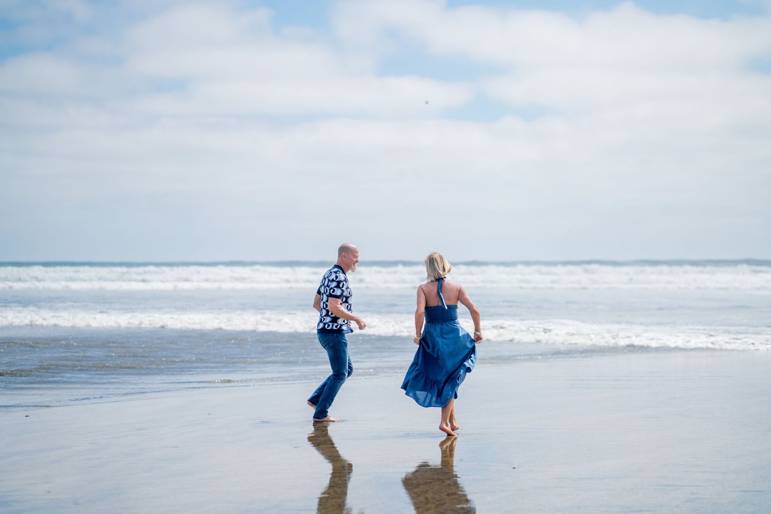 Older couple running and playing tag at low tide on the beach. Woman in blue dress, man in patterned blue and white shirt. Oceanside, CA