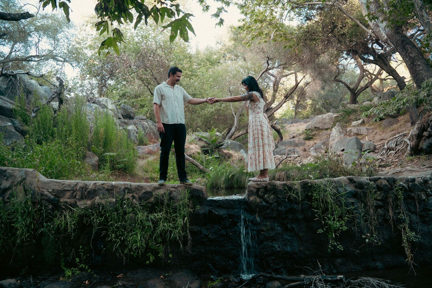 A couple holds hands on a stone bridge surrounded by lush greenery and a small waterfall in a scenic park. Vista, CA