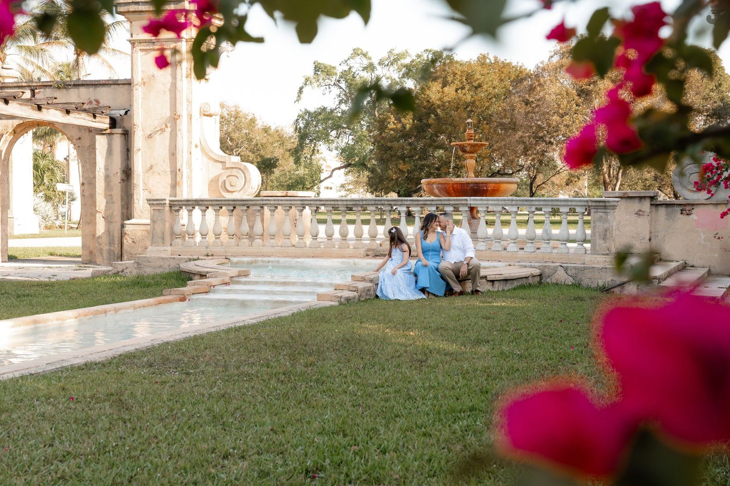 Couple in elegant attire sitting on lush garden steps near ornate stone balustrade with pink bougainvillea in foreground.
