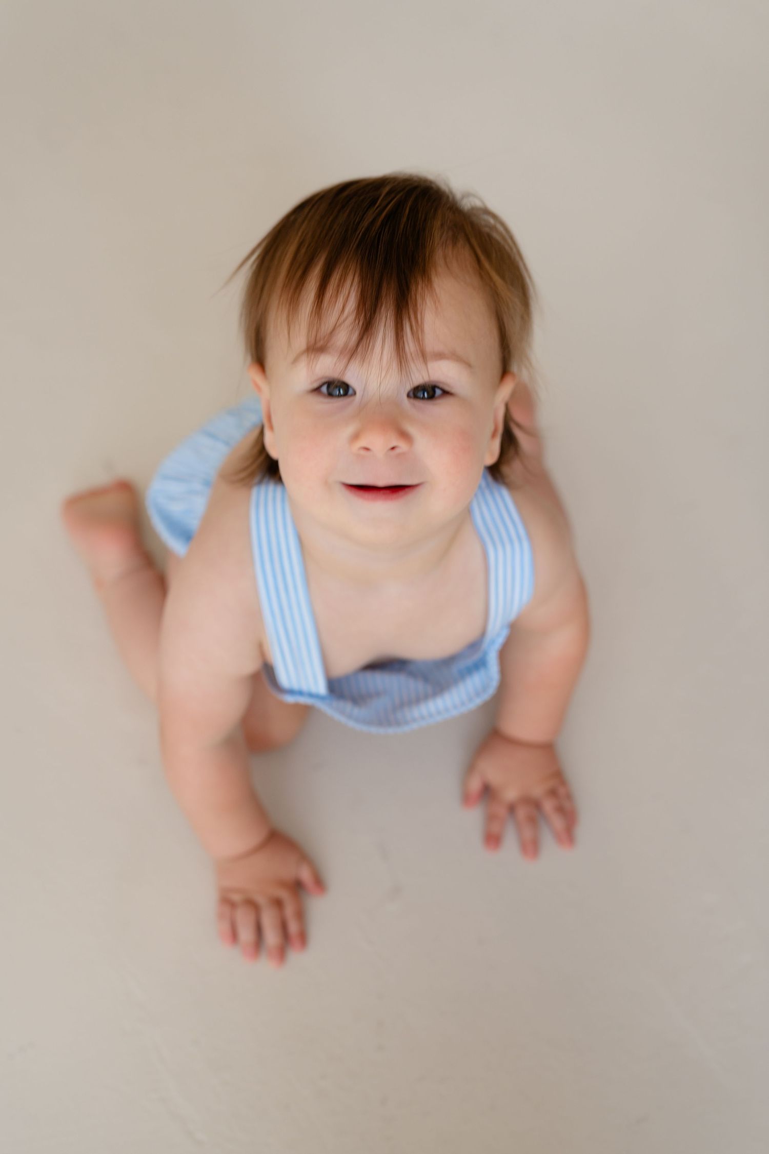 Adorable baby in blue striped romper sitting on floor, looking up with a cheerful smile and bright eyes.