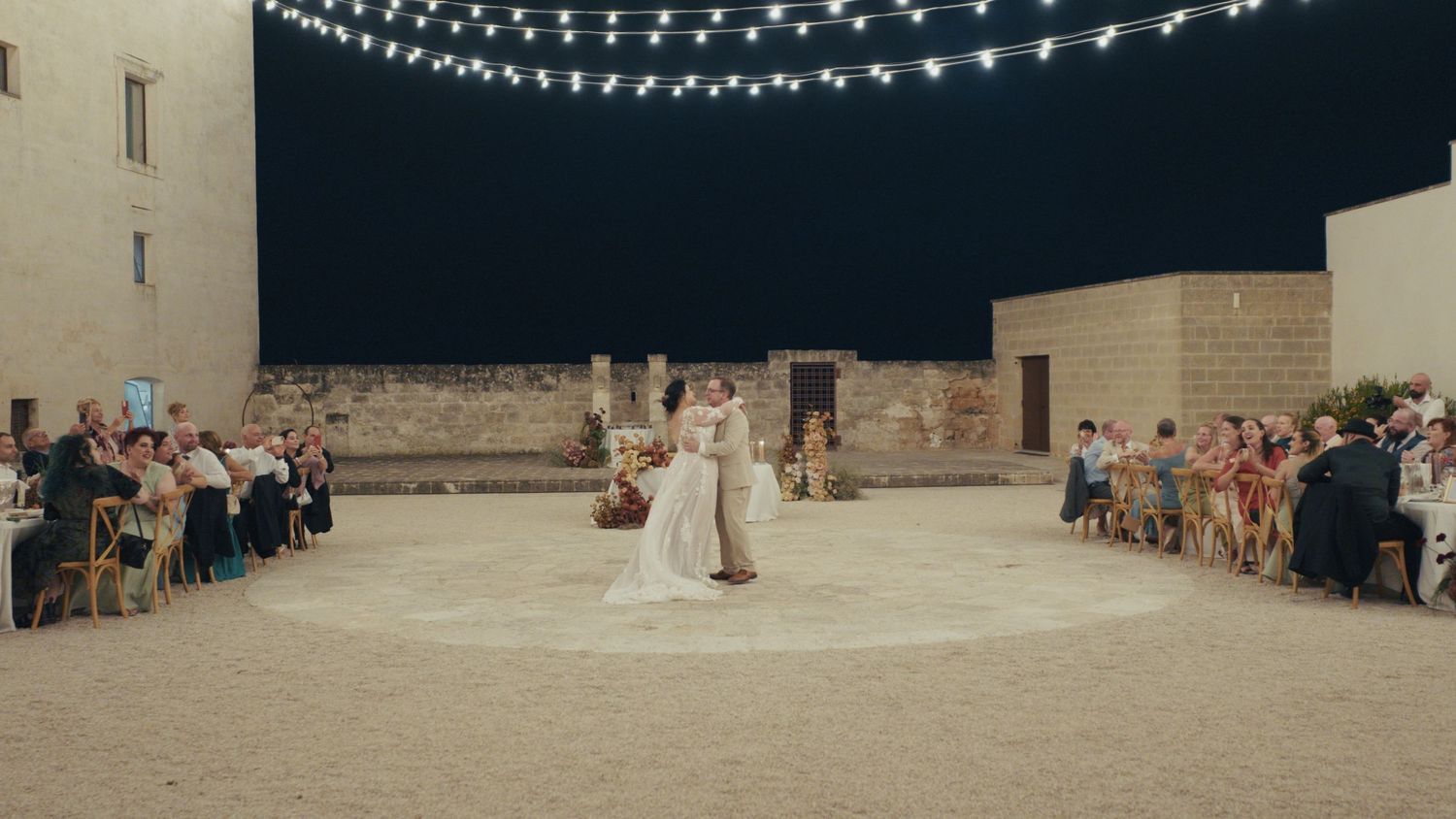 Couple shares first dance at outdoor wedding reception under string lights with guests seated around sandy courtyard at night.