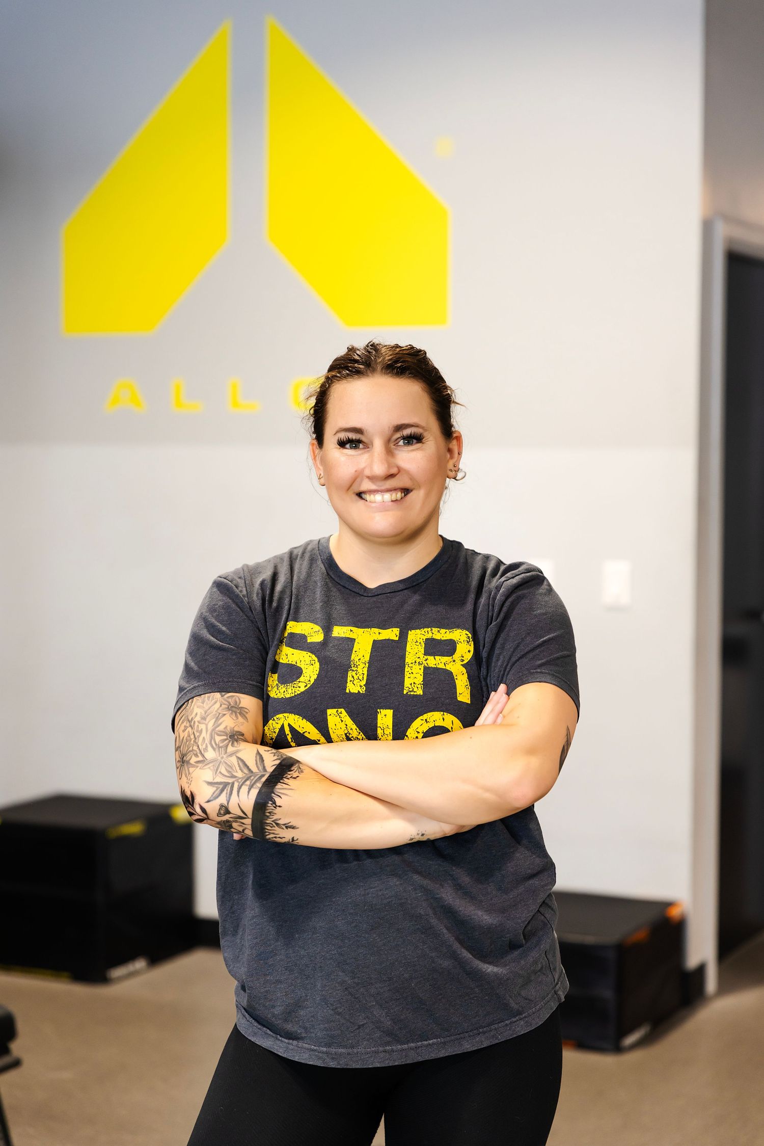Smiling fitness instructor with tattoos wearing a gray STR ONG shirt stands arms crossed in an Alloy gym.