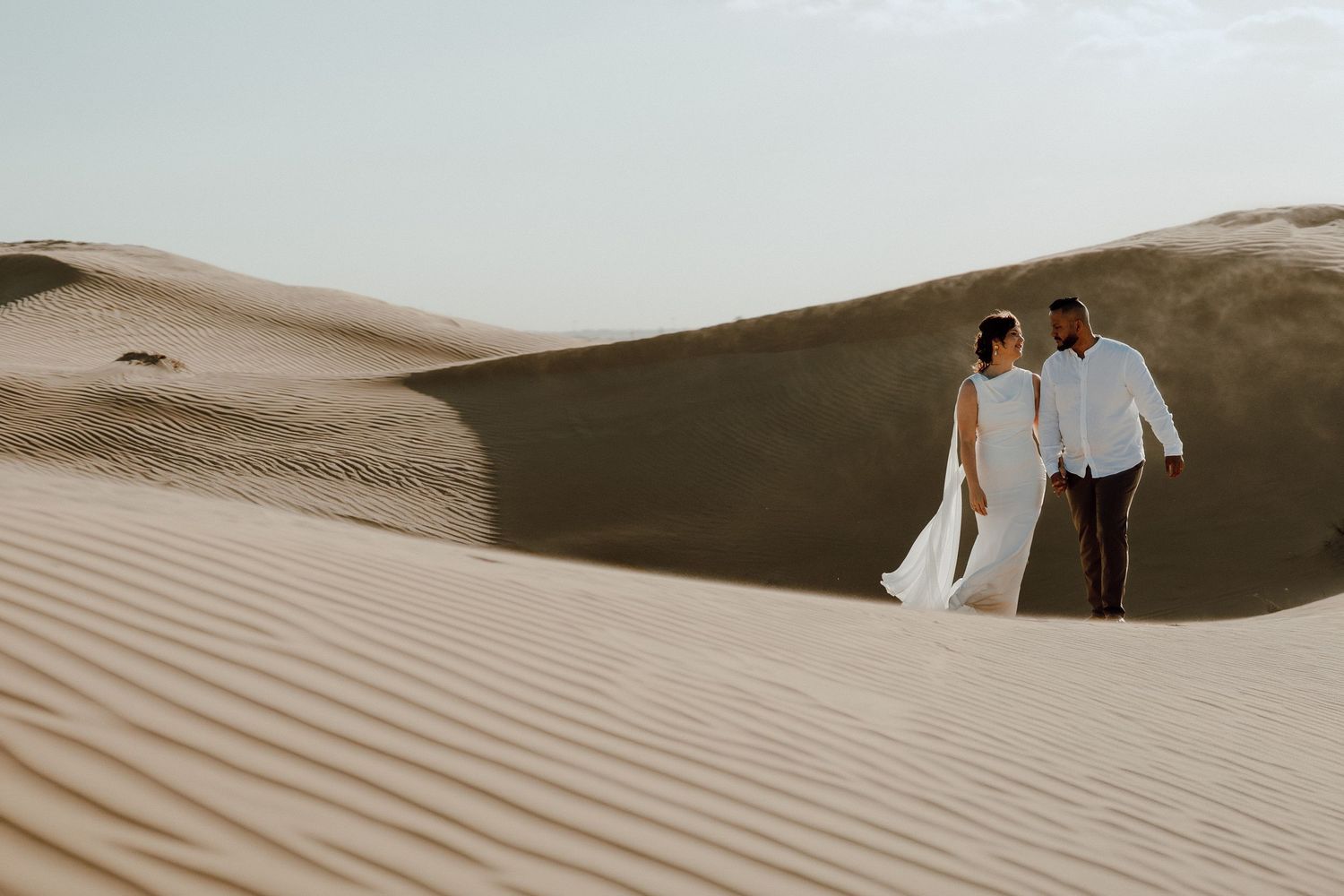 Couple in white attire walking hand in hand across golden sand dunes in a vast desert landscape.