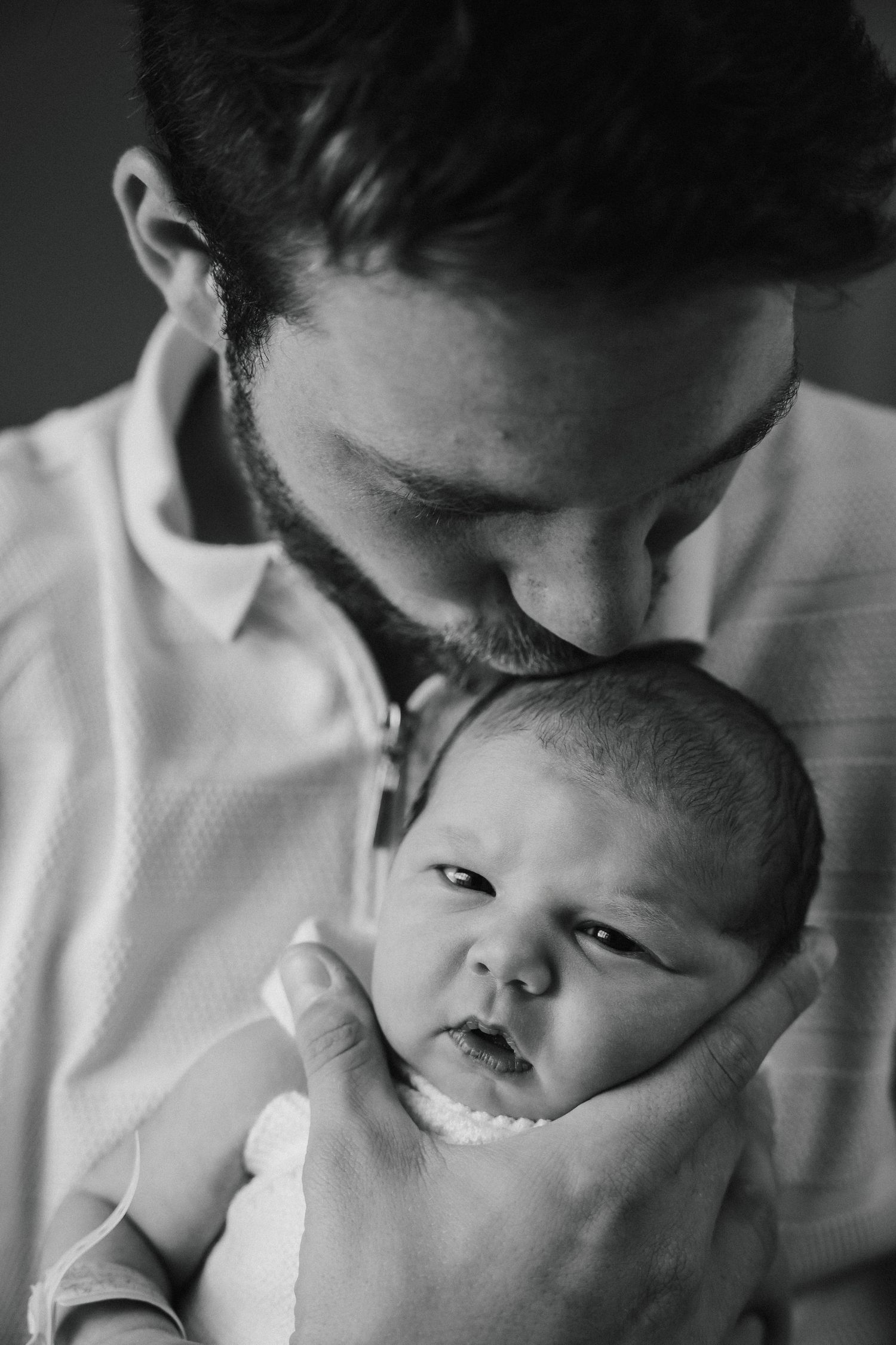 Black and white photo of a bearded father tenderly kissing his newborn baby on the forehead.