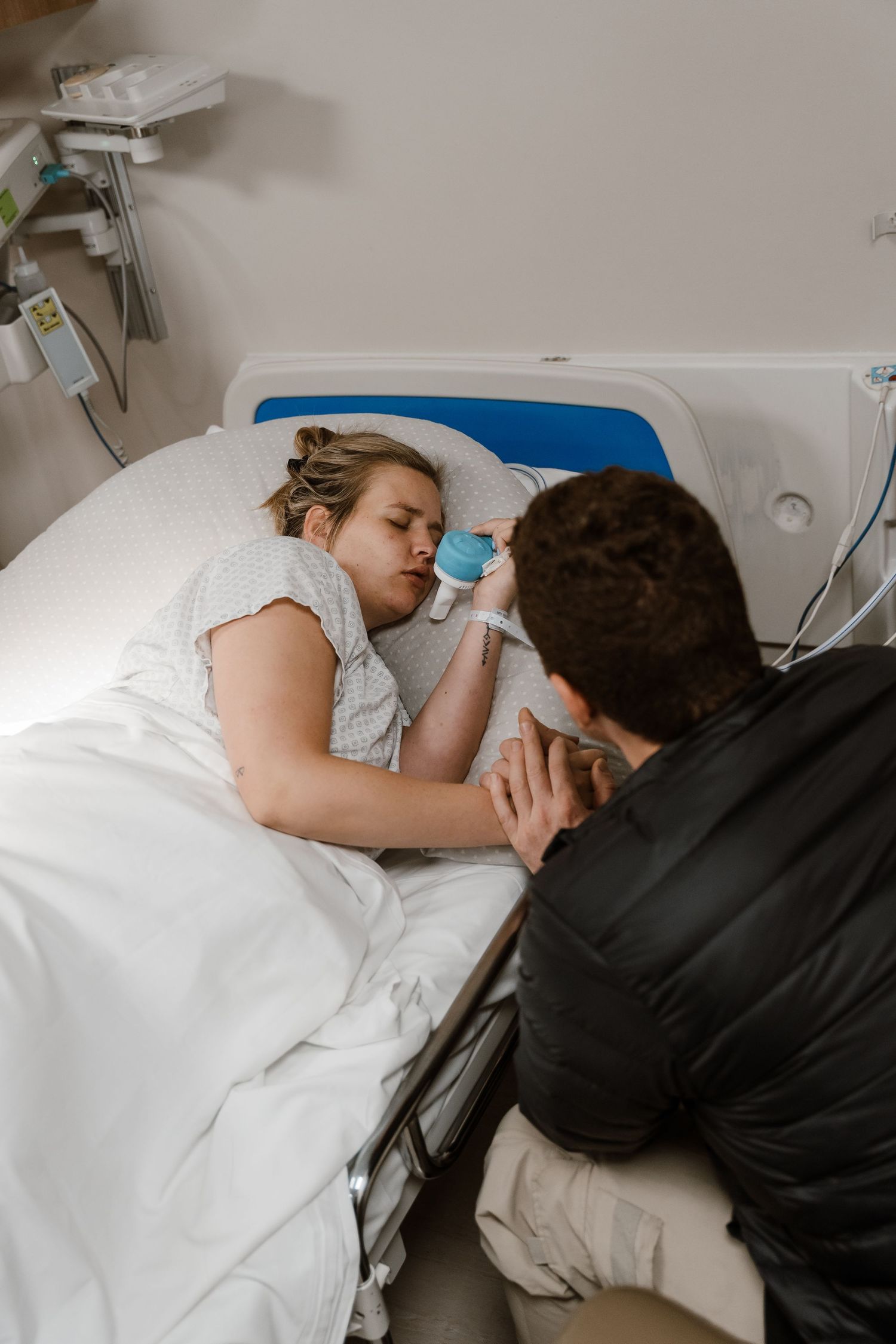 A woman in a hospital bed holds a cup while a man sits beside her, holding her hand during recovery.