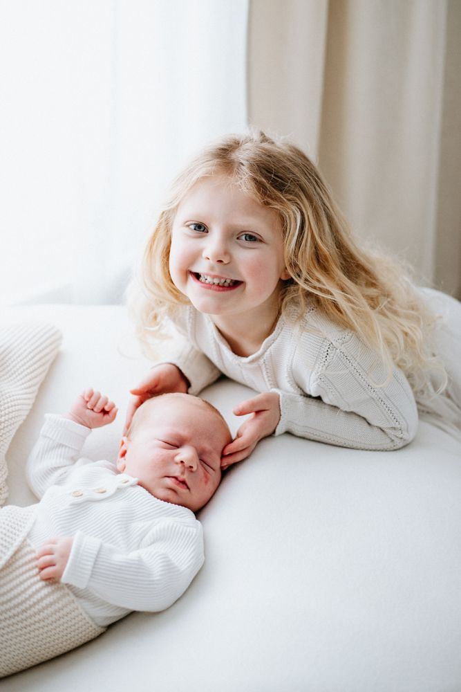 Smiling blonde girl with long hair poses with sleeping newborn baby sibling, both dressed in white on a soft surface.