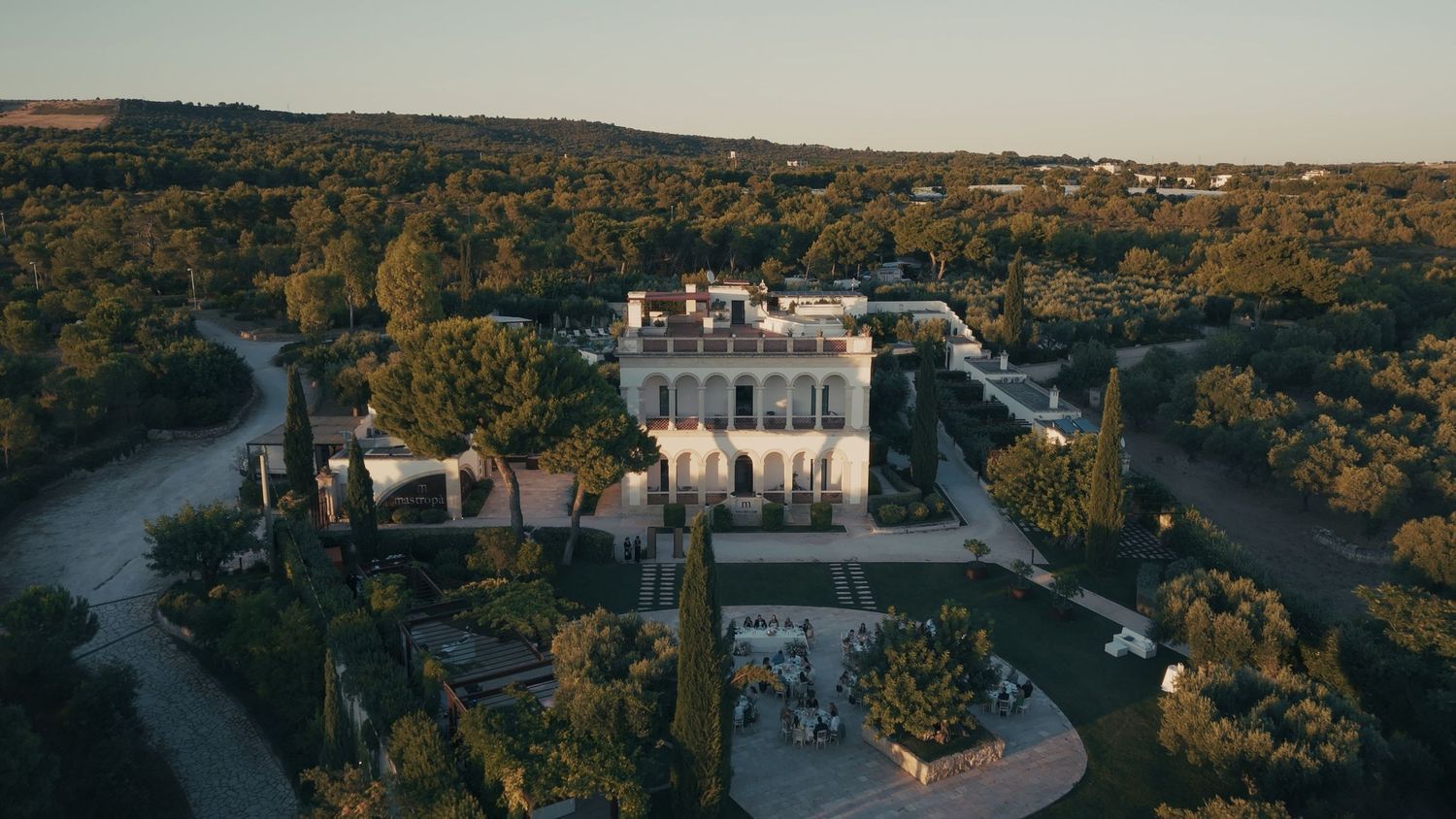 Aerial view of a grand Mediterranean villa surrounded by lush green trees at golden hour.