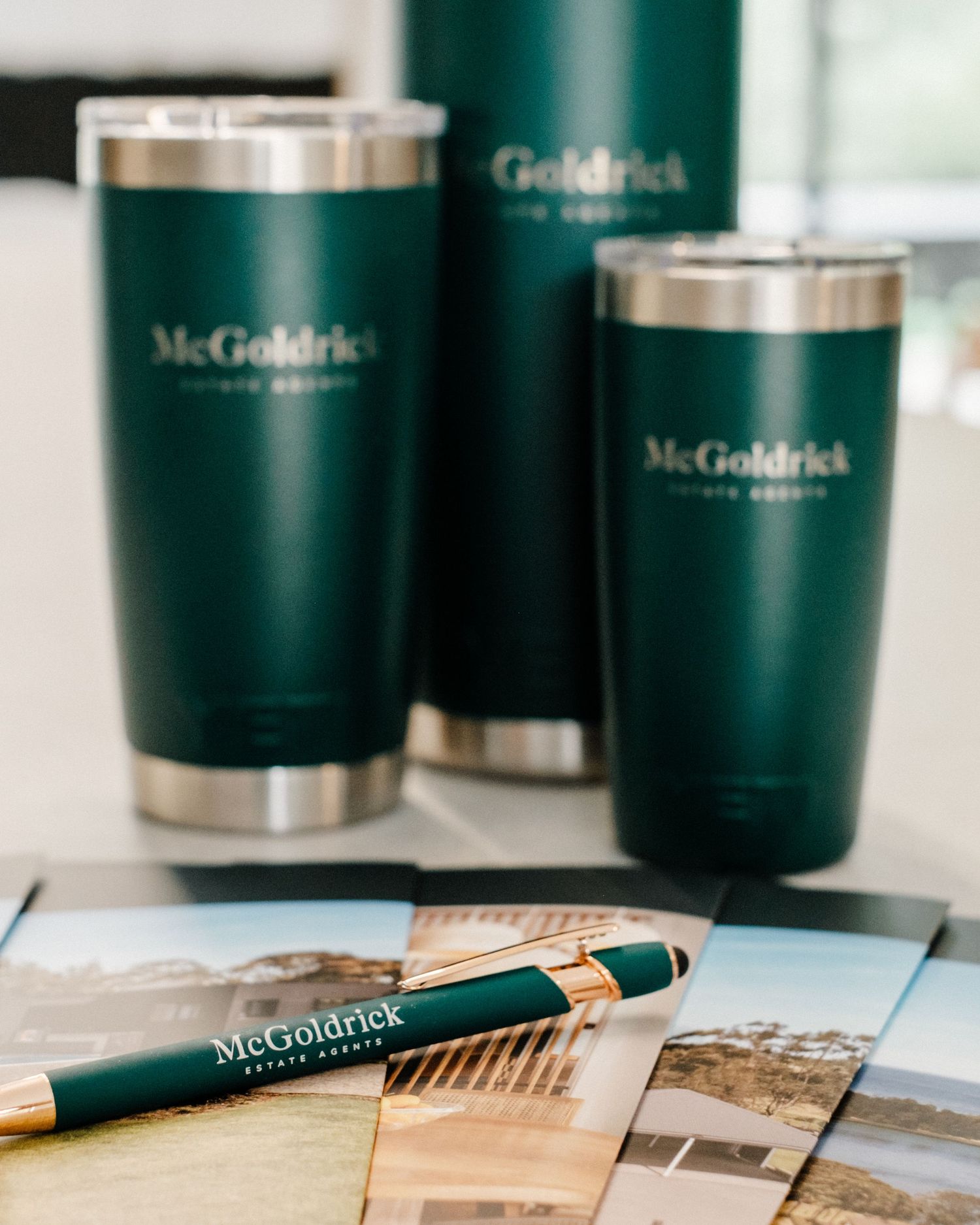Three green McGoldrick Private Property branded tumblers and a matching pen displayed with real estate brochures.