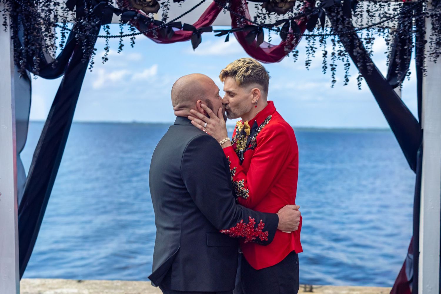 Two grooms share a kiss at their waterfront wedding ceremony under a floral arch.