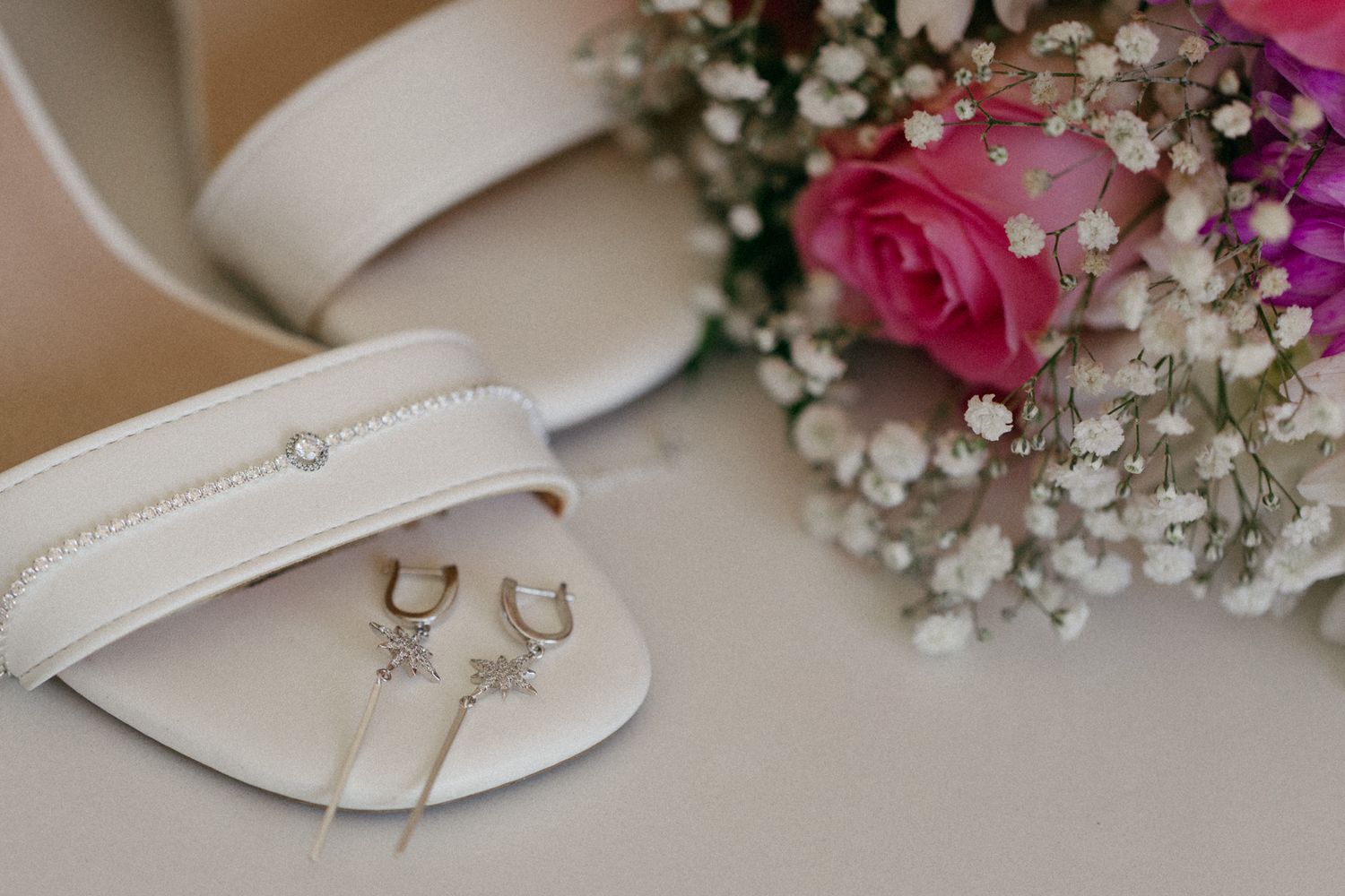 Elegant bridal flat shoes with crystal detail, gold earrings, and a pink rose bouquet with baby's breath on a neutral background.