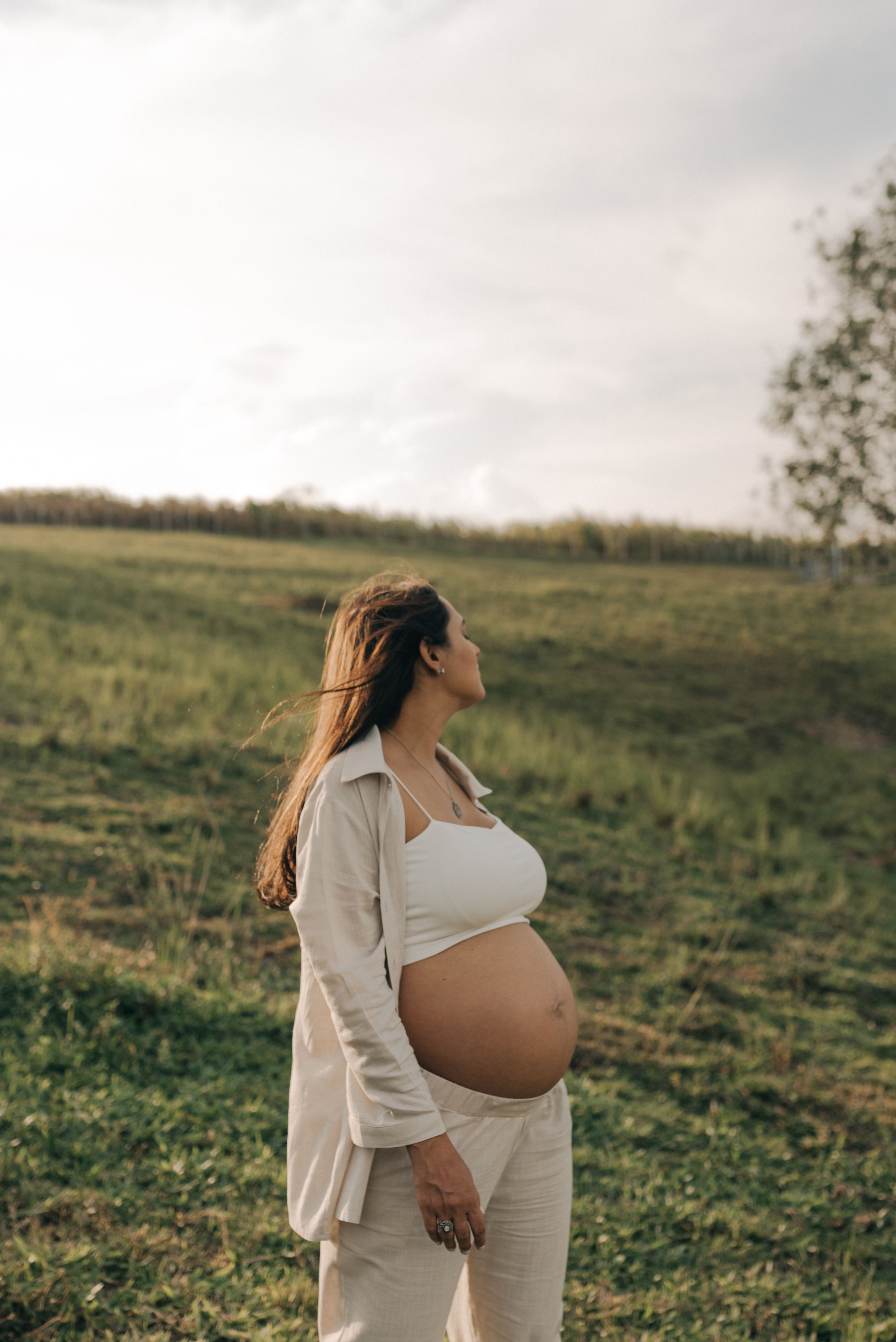Pregnant woman in white outfit standing in grassy field during cloudy day.