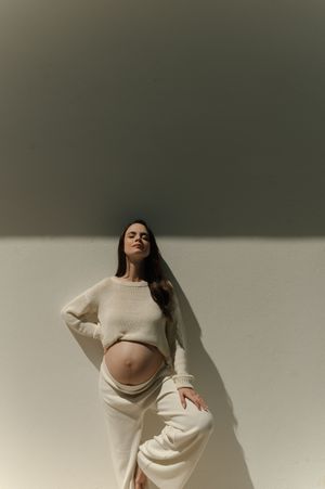A maternity portrait in cream-colored knitwear against a minimalist background with dramatic lighting and shadows.