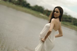 A person in a white dress stands gracefully in a natural outdoor setting near a body of water.