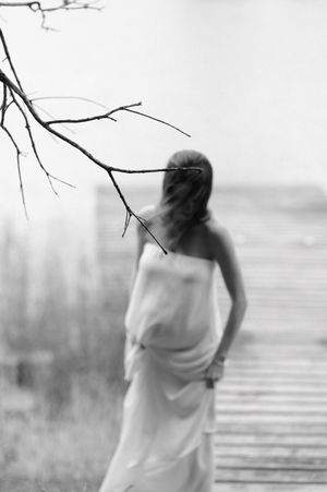 Artistic black and white photo of a figure in flowing white fabric standing on a wooden boardwalk by water.