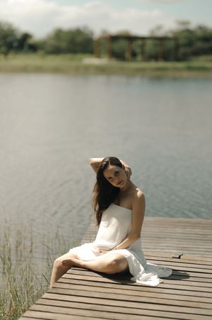 Woman in white dress lying on wooden dock by serene lake during peaceful summer afternoon.