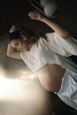 Artistic maternity portrait with dramatic lighting shows a pregnant woman in white clothing against a dark background.
