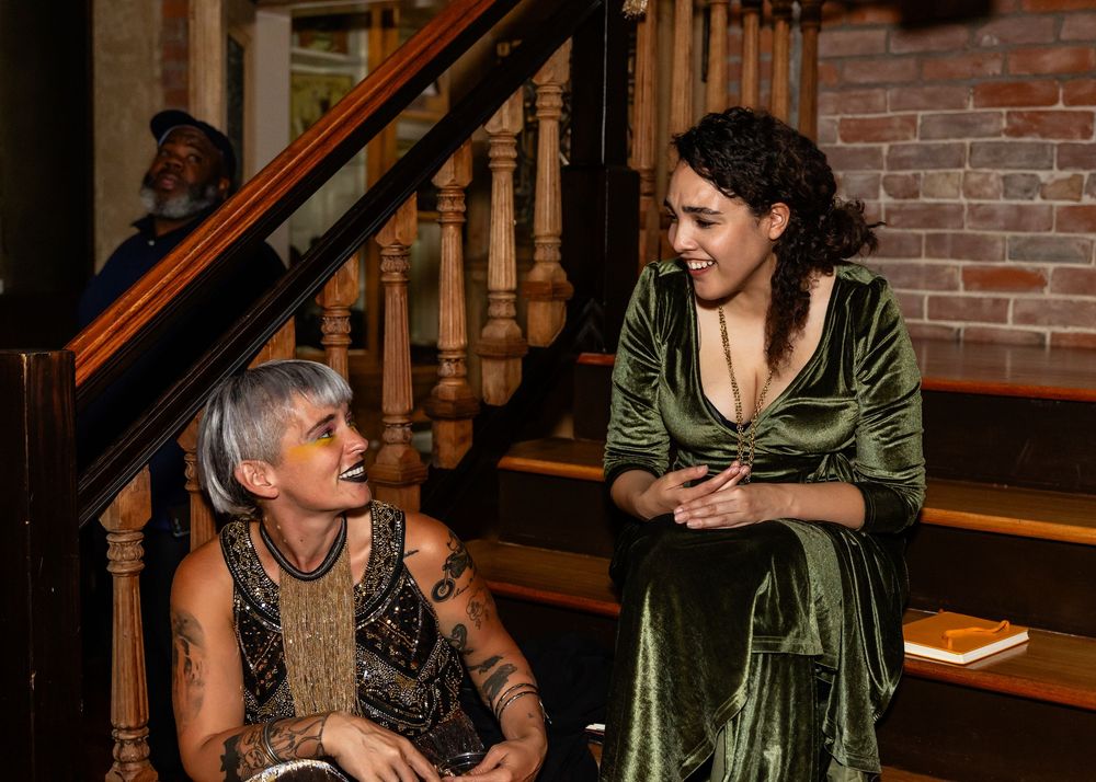 Two women in elegant attire chat warmly near a staircase in a warmly lit venue.