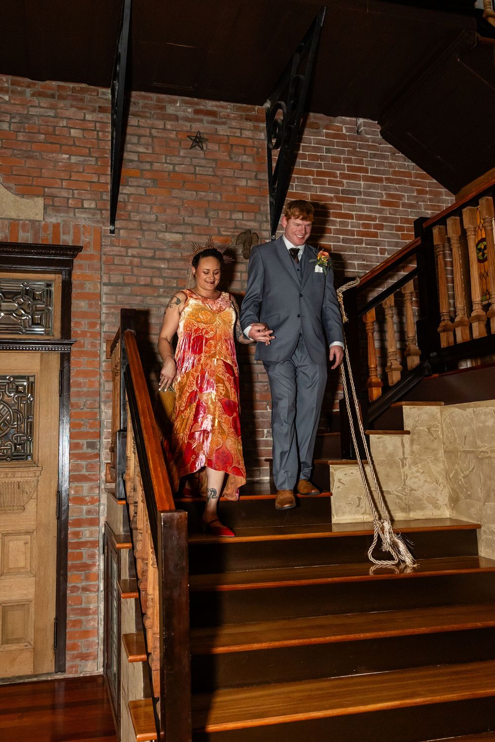 Couple descending wooden staircase in brick venue, woman in red dress and man in gray suit at wedding reception.