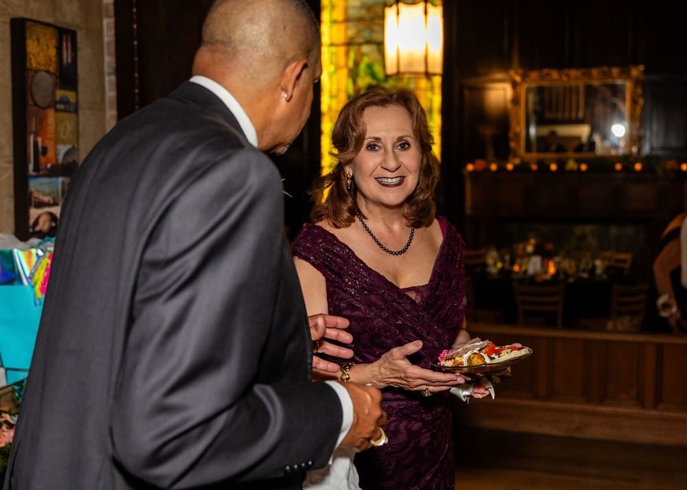 Woman in burgundy dress holding a plate at an elegant indoor event, conversing with a man in a gray suit.