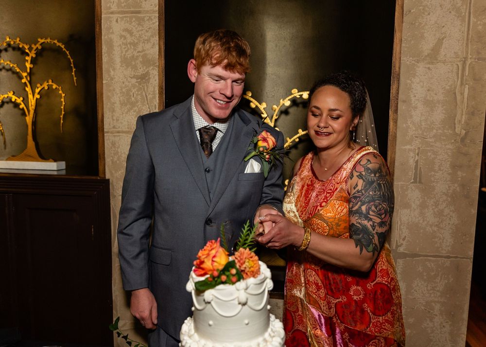 Bride and groom cutting white wedding cake together, bride in colorful sari, groom in gray suit.