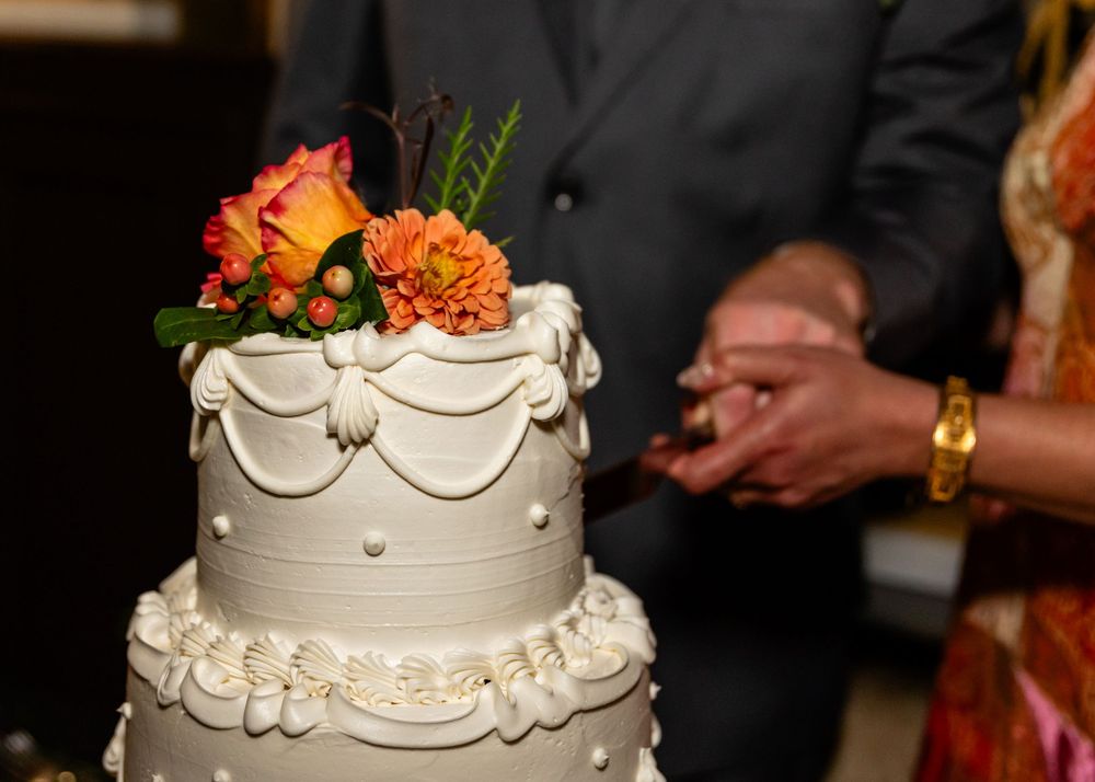 Elegant white three-tier wedding cake topped with orange and pink flowers, with couple cutting cake in background.