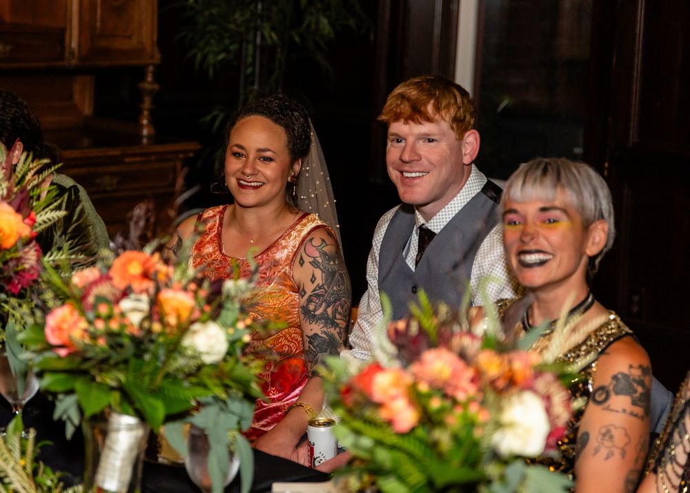 Three smiling wedding guests seated at a floral-decorated reception table with colorful bouquets in a warmly lit venue.