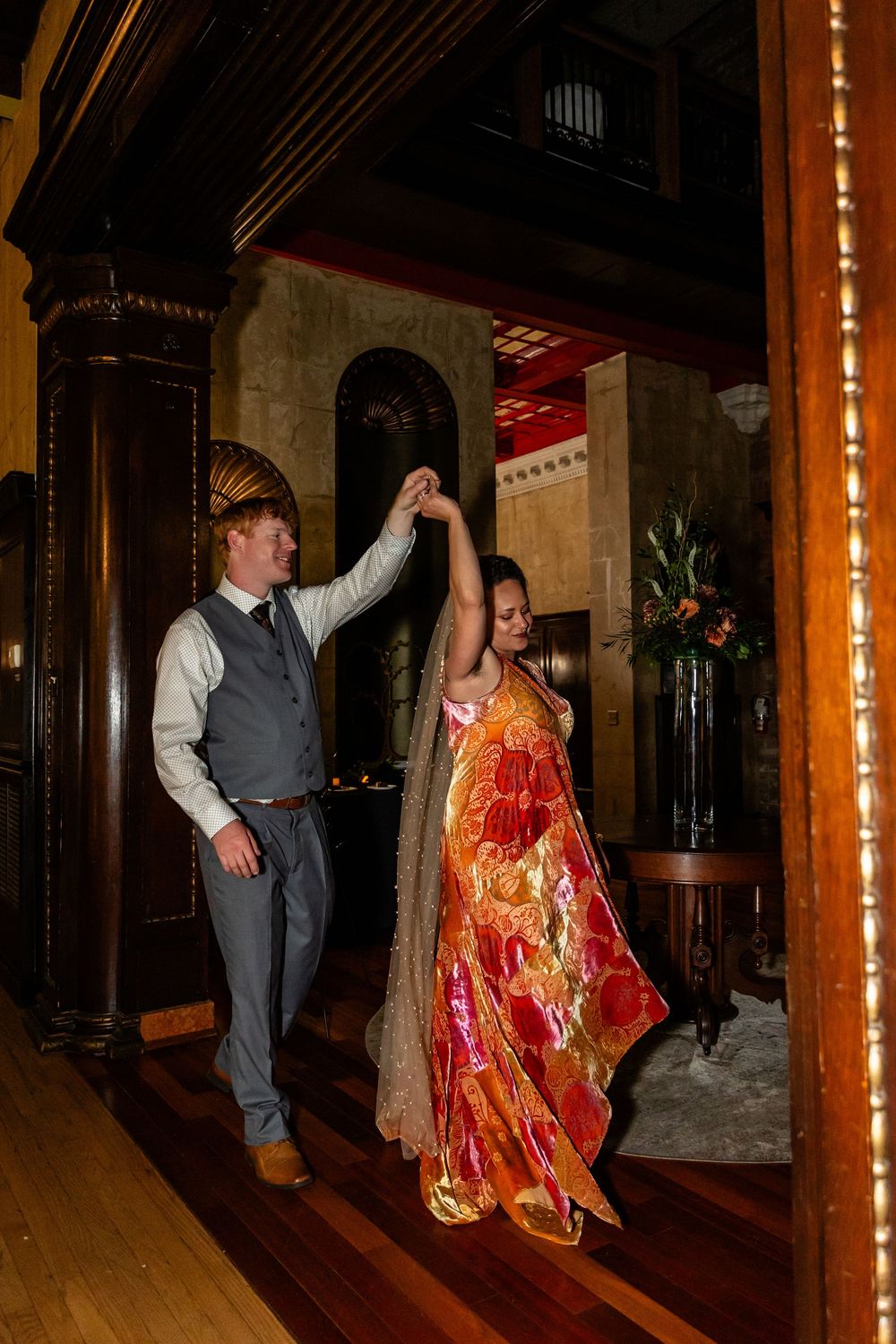 Couple dancing elegantly in a grand wooden hall, man in gray vest twirling woman in vibrant orange floral gown.