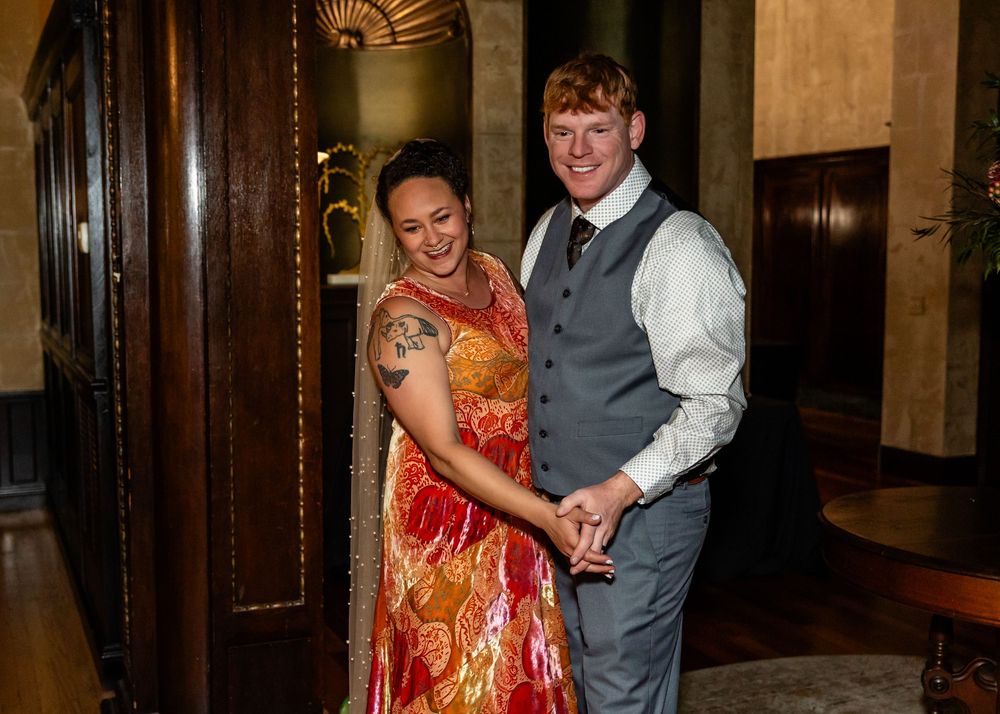 Smiling couple holding hands in elegant venue, woman in red floral dress with tattoo, man in gray vest and tie.