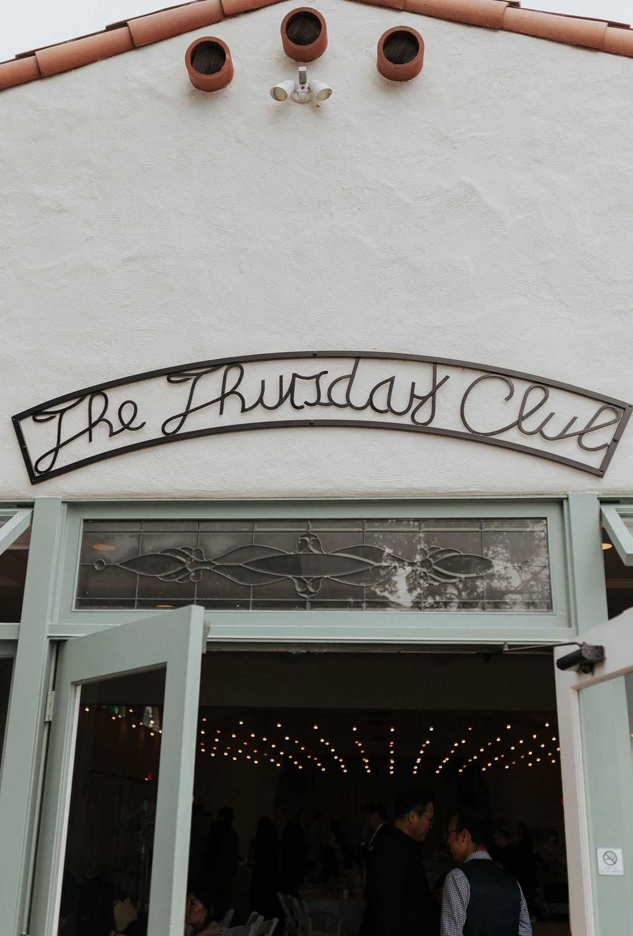 The Thursday Club venue entrance with mint green doors, string lights, and metal script sign on white stucco wall.