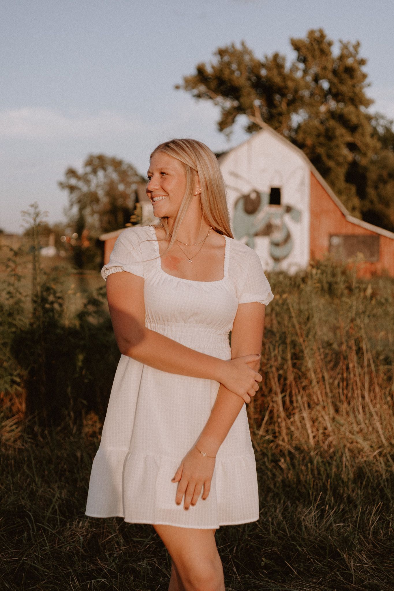 Blonde woman in white puff sleeve mini dress laughing outdoors at golden hour near a barn.