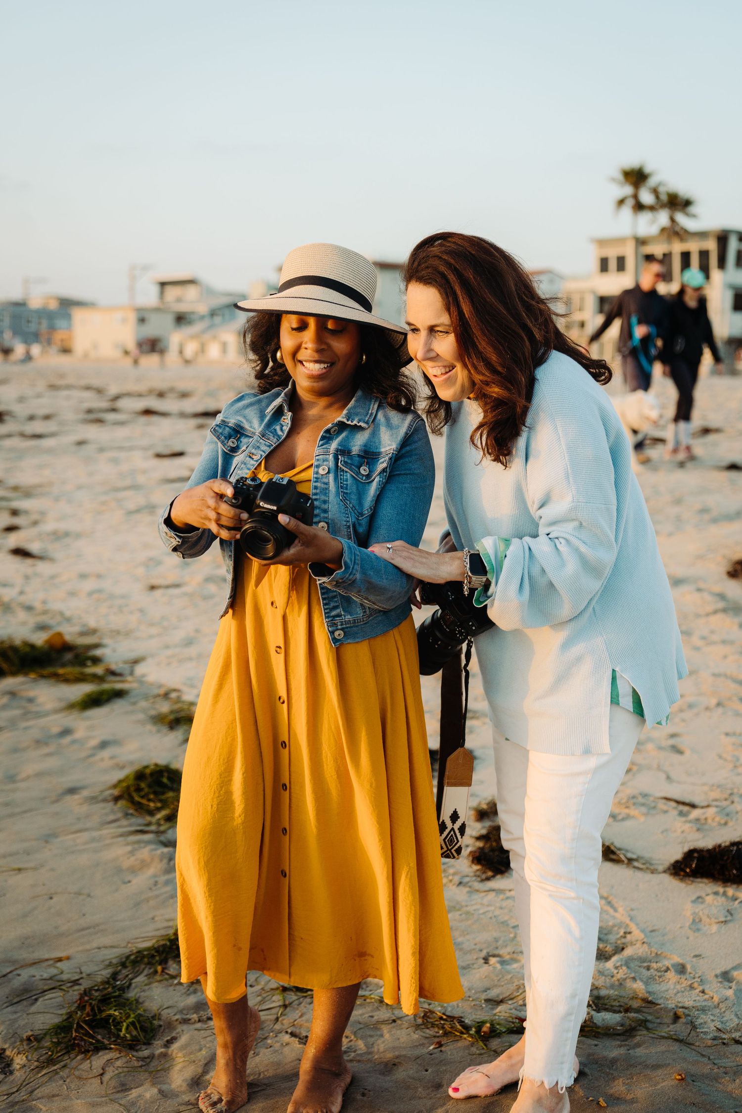 Two women smiling at a camera on a sunny beach, one in a yellow dress and hat, the other in a light blue outfit.
