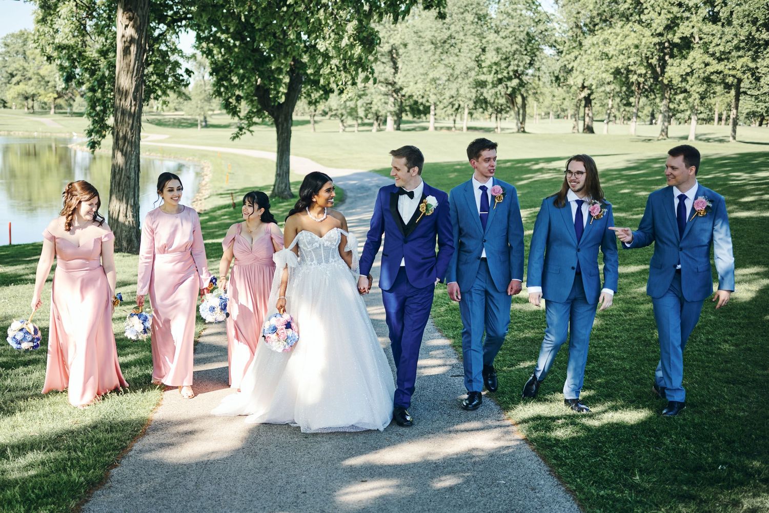Wedding party walks outdoors near a pond, bridesmaids in pink dresses, groomsmen in blue suits, bride in white gown.