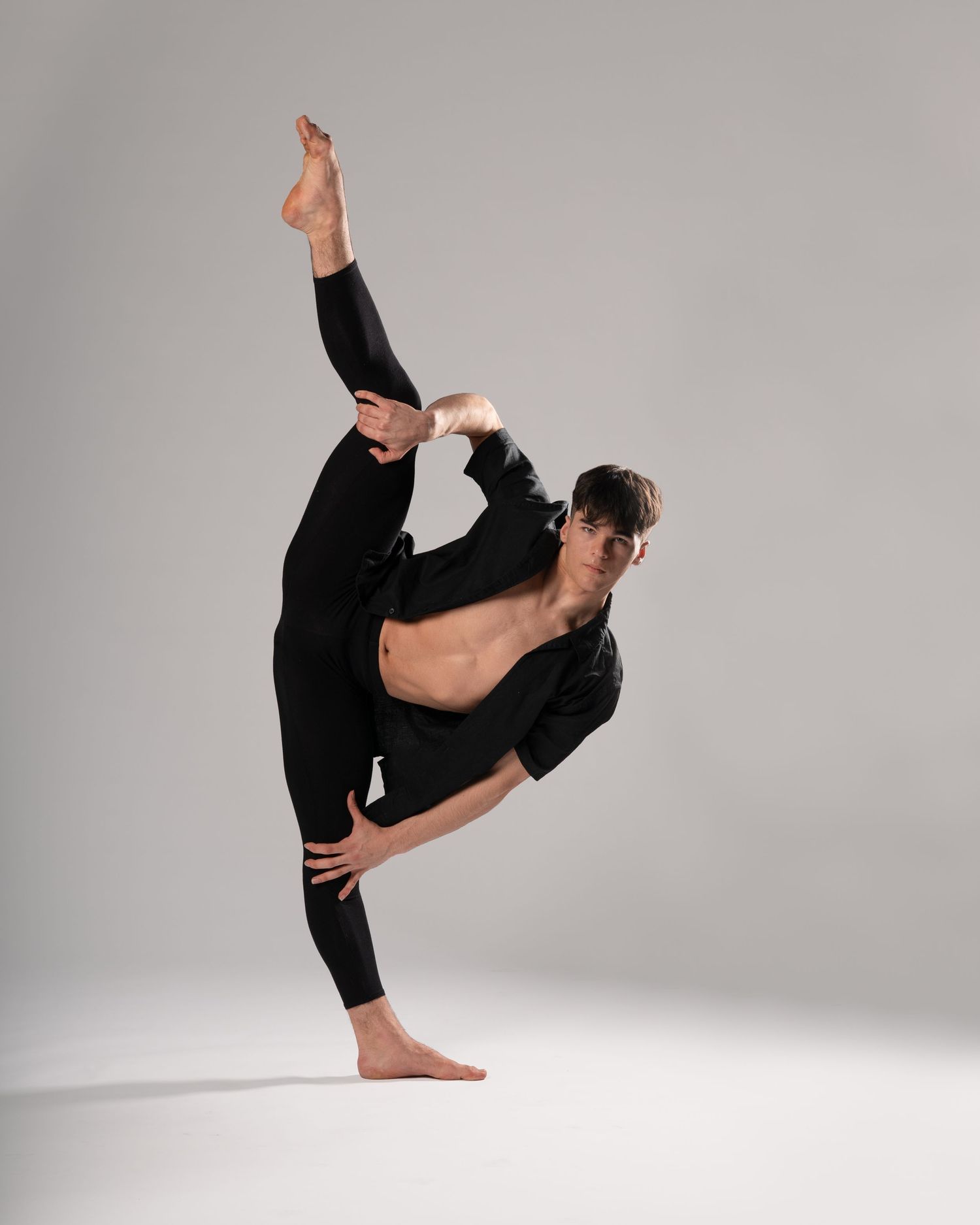 A dancer in a flowing black skirt and red scarf performs an elegant pose with arms raised overhead in a studio.