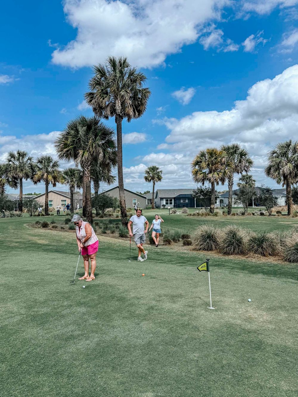 Golfers playing on a lush green golf course surrounded by tall palm trees and residential homes under a partly cloudy sky.